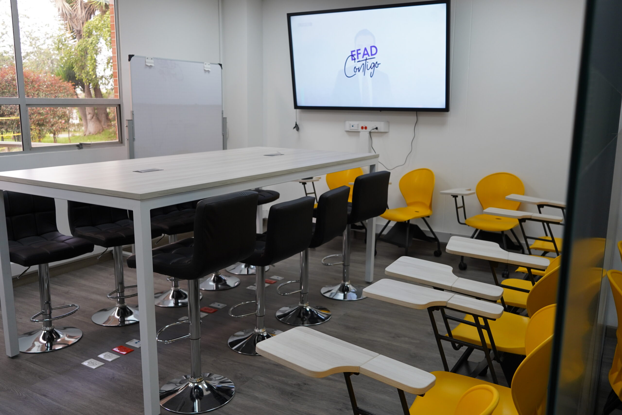 Sala de formación con mesa alta, sillas amarillas y pantalla con el logo «EFAD Contigo»