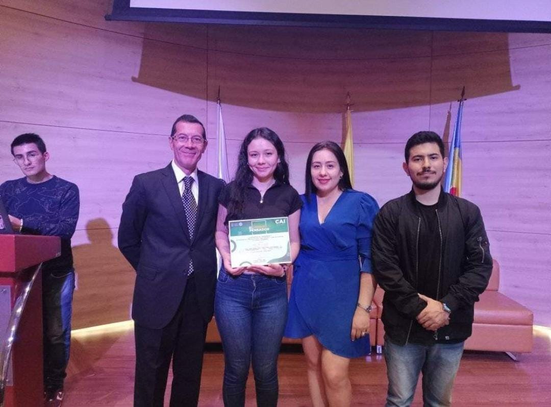 Una estudiante recibe su certificado de los Premios El Pensador IPA 2024 en el auditorio, acompañada por tres docentes.